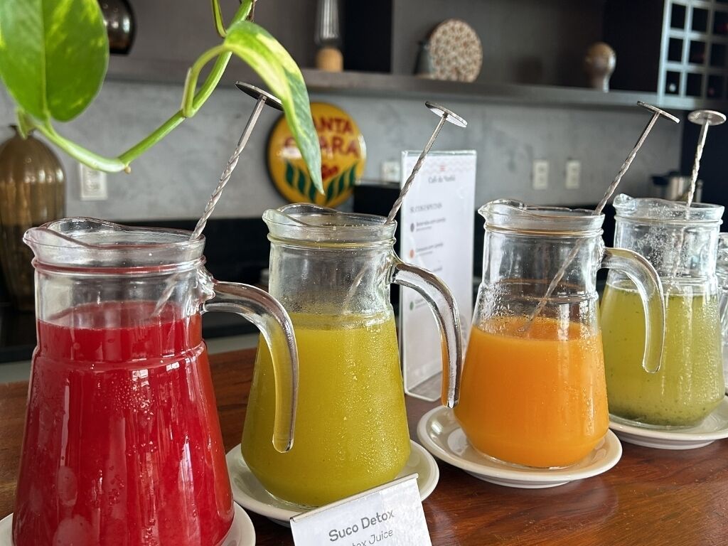 Vier glazen karafjes met kleurrijke drankjes op een tafel, met een bordje waarop 'Suco Detox' staat.