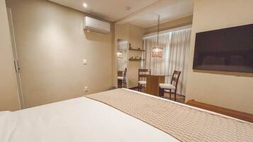 A hotel room with a large bed, a desk with chairs, a window with curtains, and an air conditioning unit.