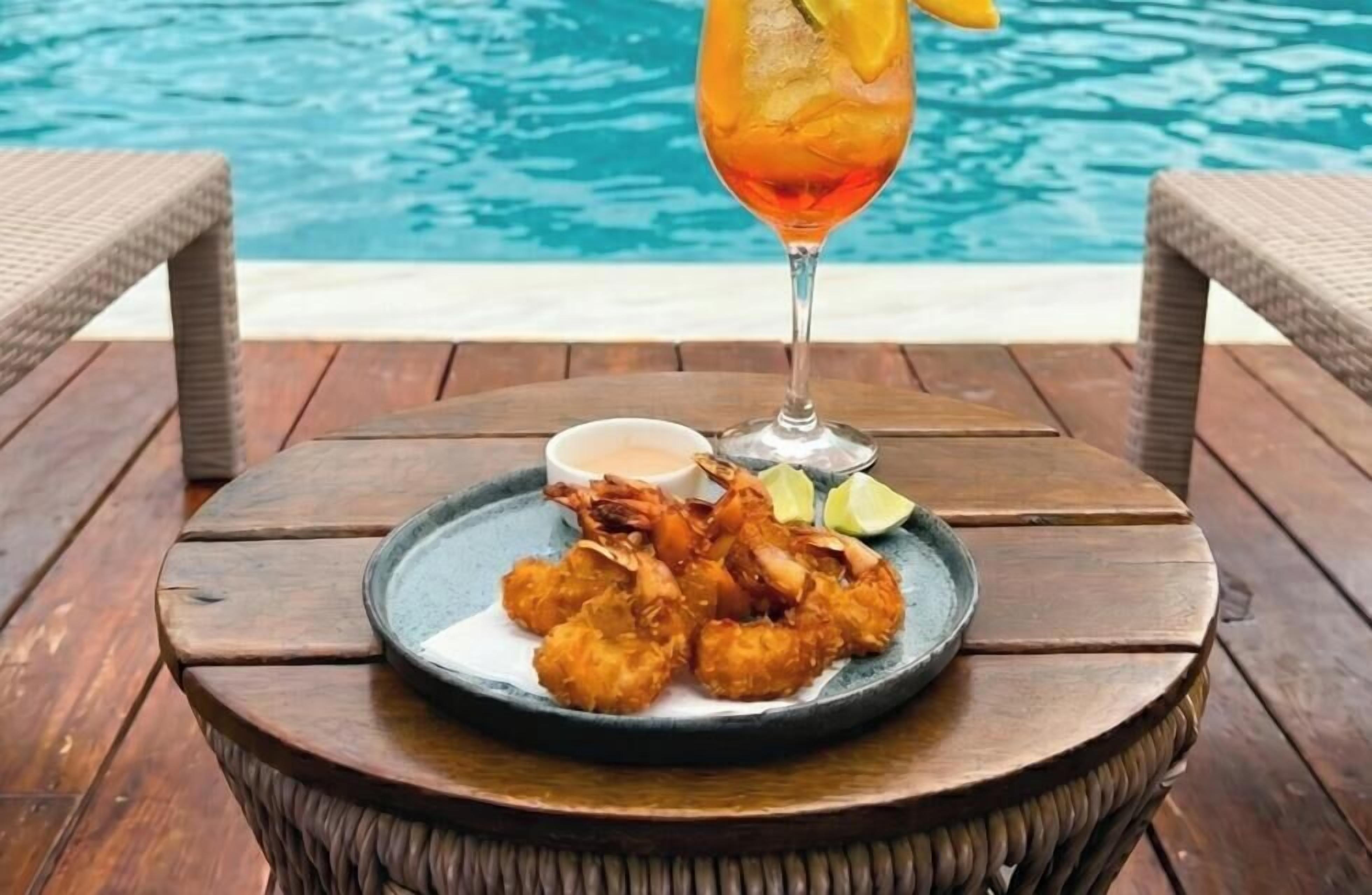 Una mesa de madera con un plato de camarones fritos y un vaso de bebida junto a una piscina.