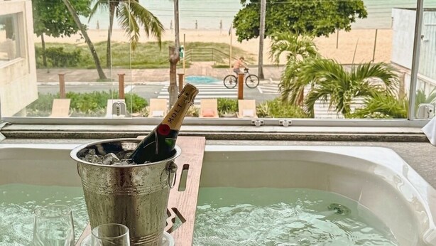 A hot tub with a champagne bottle and glasses, a wooden tray, and a view of a beach and palm trees.
