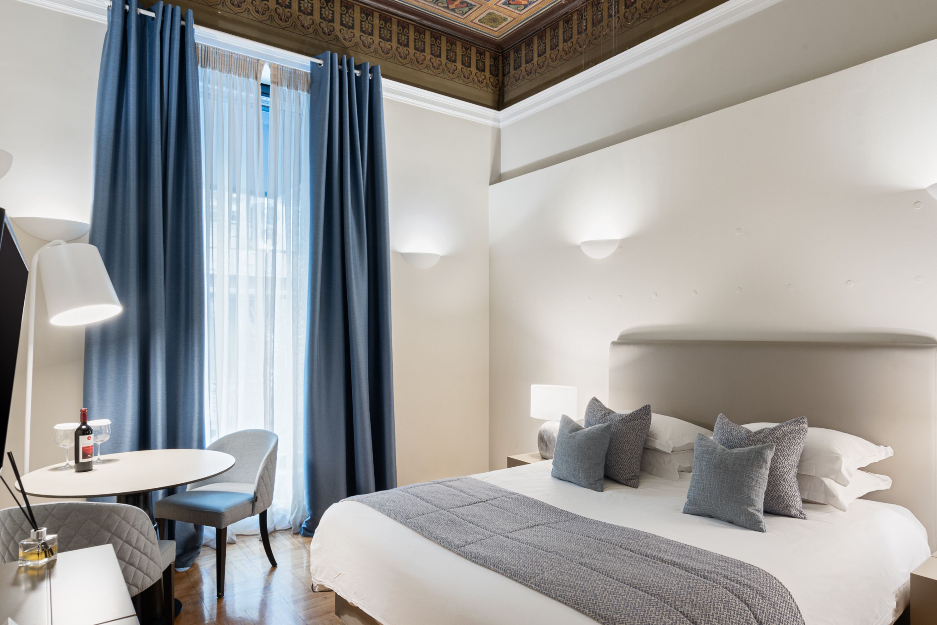 A hotel room with a bed, bedside lamps, a table with a chair, and a window with curtains.