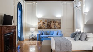 Ein geräumiges Zimmer mit einem großen Bett, einer Couch und einem Kamin. Die Decke weist aufwendige Verzierungen und ein großes Gemälde auf.