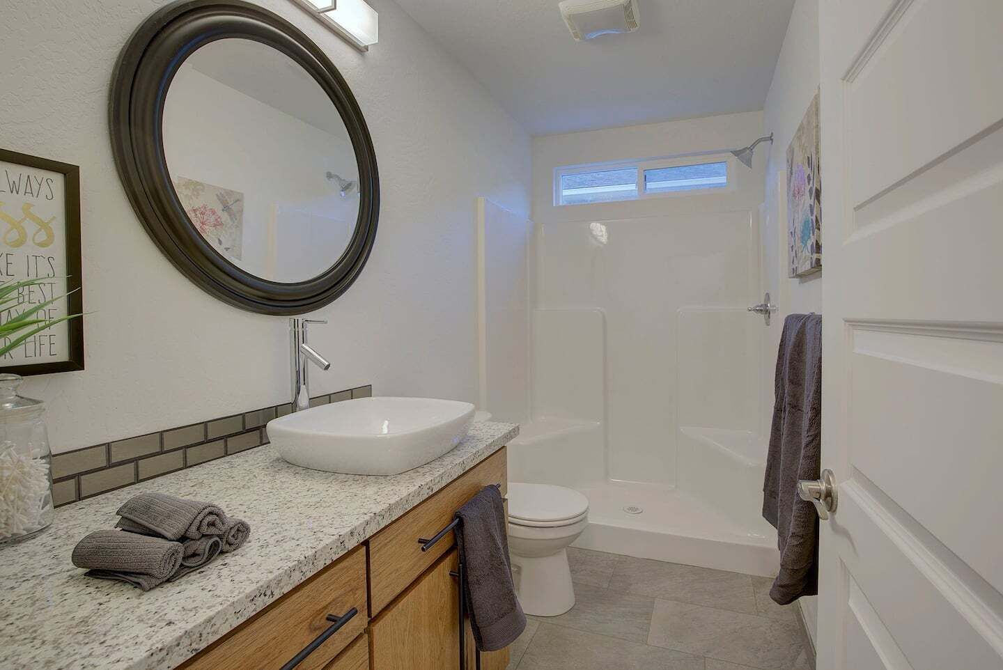Une salle de bain avec un miroir rond, un lavabo, des toilettes et une douche. Il y a des serviettes sur le comptoir et une citation encadrée au mur.