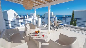 A terrace with white furniture, a table with fruit and drinks, and a view of the sea and buildings.