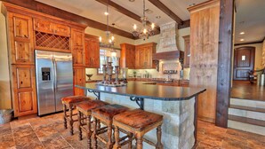 A spacious kitchen with wooden cabinets, a large island with stools, a stainless steel refrigerator, and a stone fireplace.