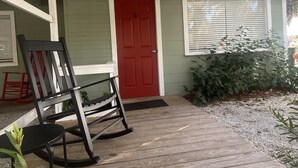 A wooden porch with a rocking chair, a red door, and a window with blinds.