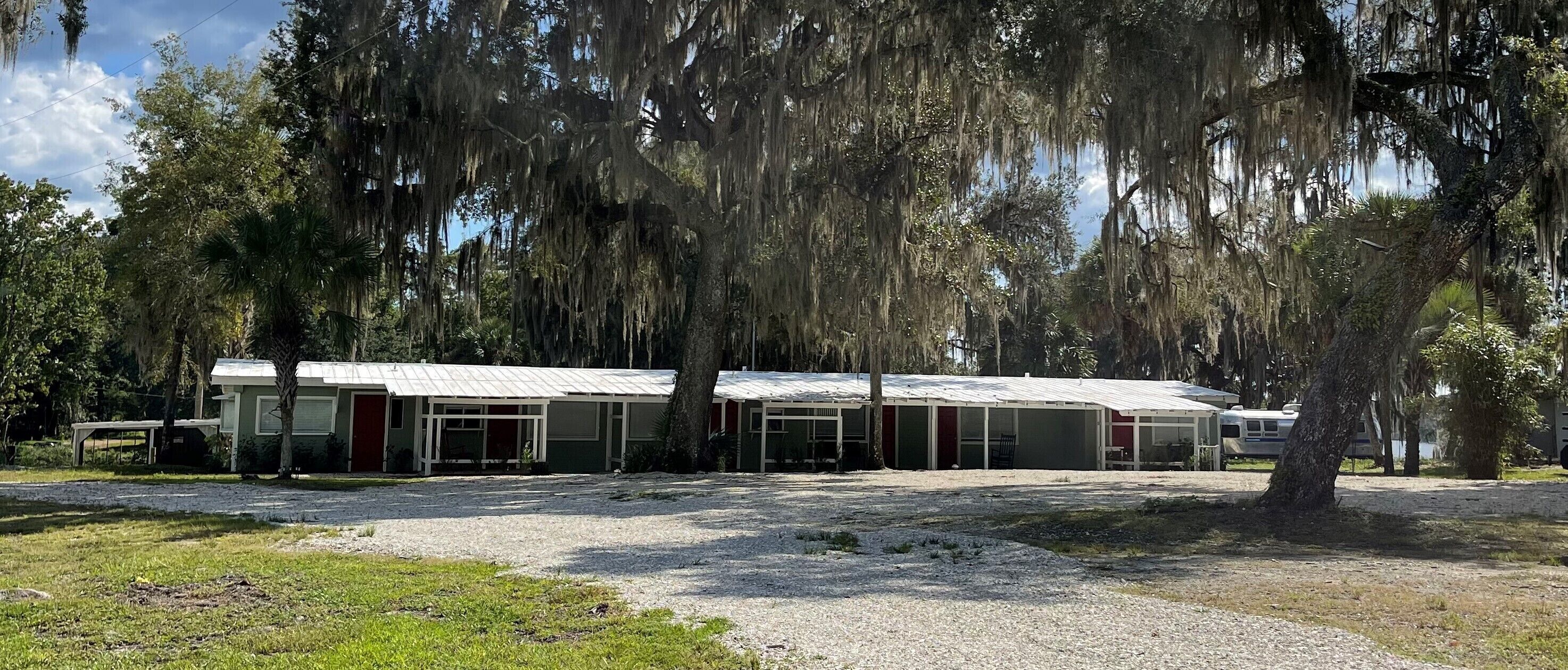 A row of motel buildings with a gravel parking area and trees with hanging moss.
