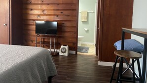 A room with a wooden wall, a bed, a TV, a fan, and a bathroom visible through an open door.