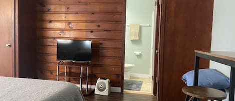 Ein Zimmer mit Holzwand, einem Bett, einem Fernseher, einem Ventilator und einem durchgegangene Tür erkennbaren Badezimmer.