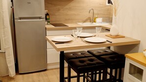 Een compacte keuken met witte kasten, een roestvrijstalen koelkast, een houten aanrechtblad en een eethoek met een tafel en twee stoelen.