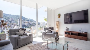 A modern living room with a sofa, armchair, and coffee table, a TV mounted on the wall, and a view of a cityscape through large windows. - Penthouse 1BR + Rooftop Paradise | Romantic Zone (Puerto Vallarta)