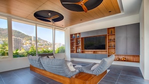 A modern living room with a large window, built-in entertainment unit, and a wooden ceiling fan. - Penthouse 1BR + Rooftop Paradise | Romantic Zone (Puerto Vallarta)