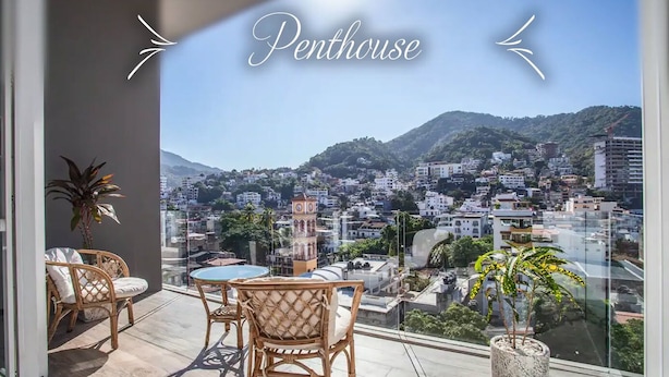 A balcony with wicker furniture, a potted plant, and a view of a cityscape with a clock tower. - Penthouse 1BR + Rooftop Paradise | Romantic Zone (Puerto Vallarta)