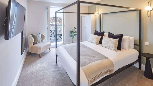 Une chambre moderne avec un lit à baldaquin, un téléviseur à écran plat fixé au mur, un petit coin salon avec une chaise et une table, et un balcon offrant une vue sur la ville.
