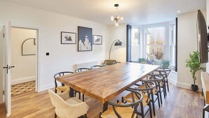 Un coin repas avec une table en bois, six chaises et un lustre. Des tableaux encadrés ornent le mur, ainsi qu’un radiateur et une porte au motif circulaire.