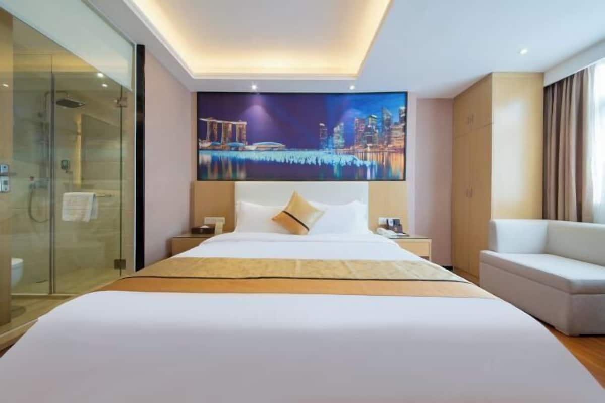 A hotel room with a large bed, a glass shower enclosure, a cityscape painting on the wall, and a white sofa.