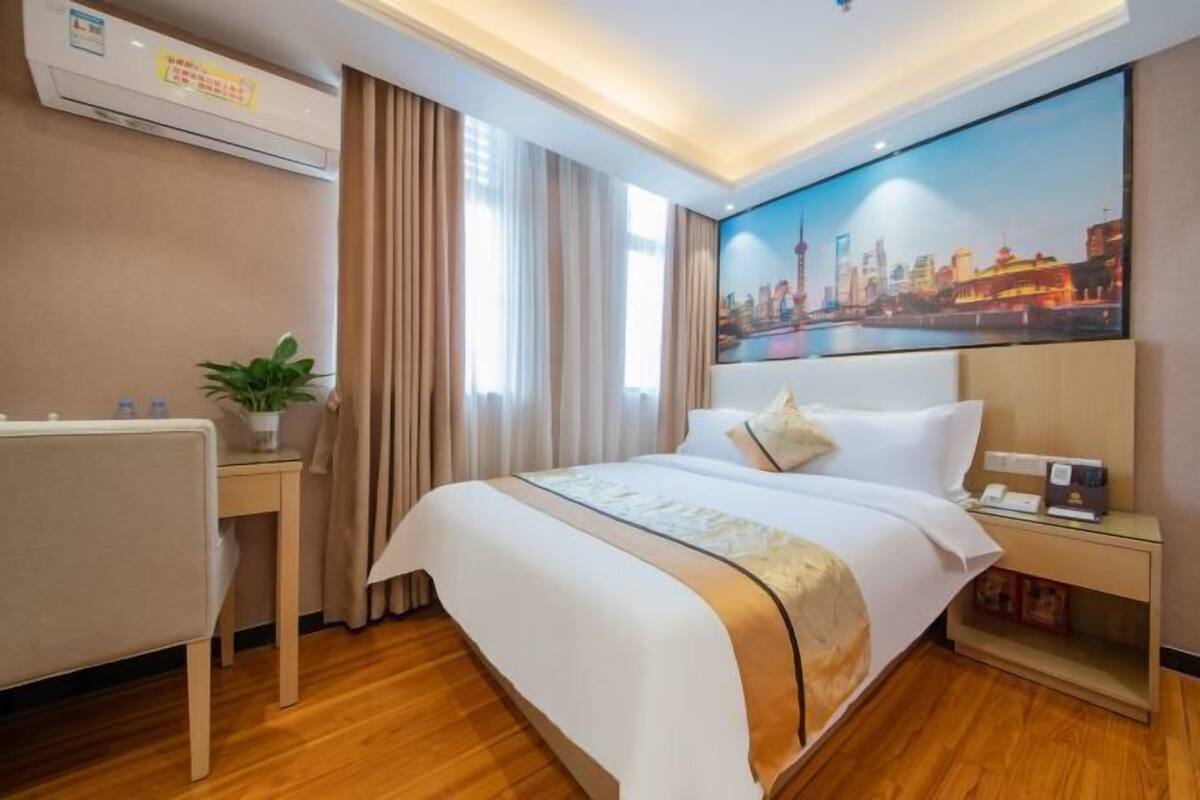 A hotel room with a large bed, a desk, a nightstand, a wall-mounted air conditioner, and a cityscape painting.