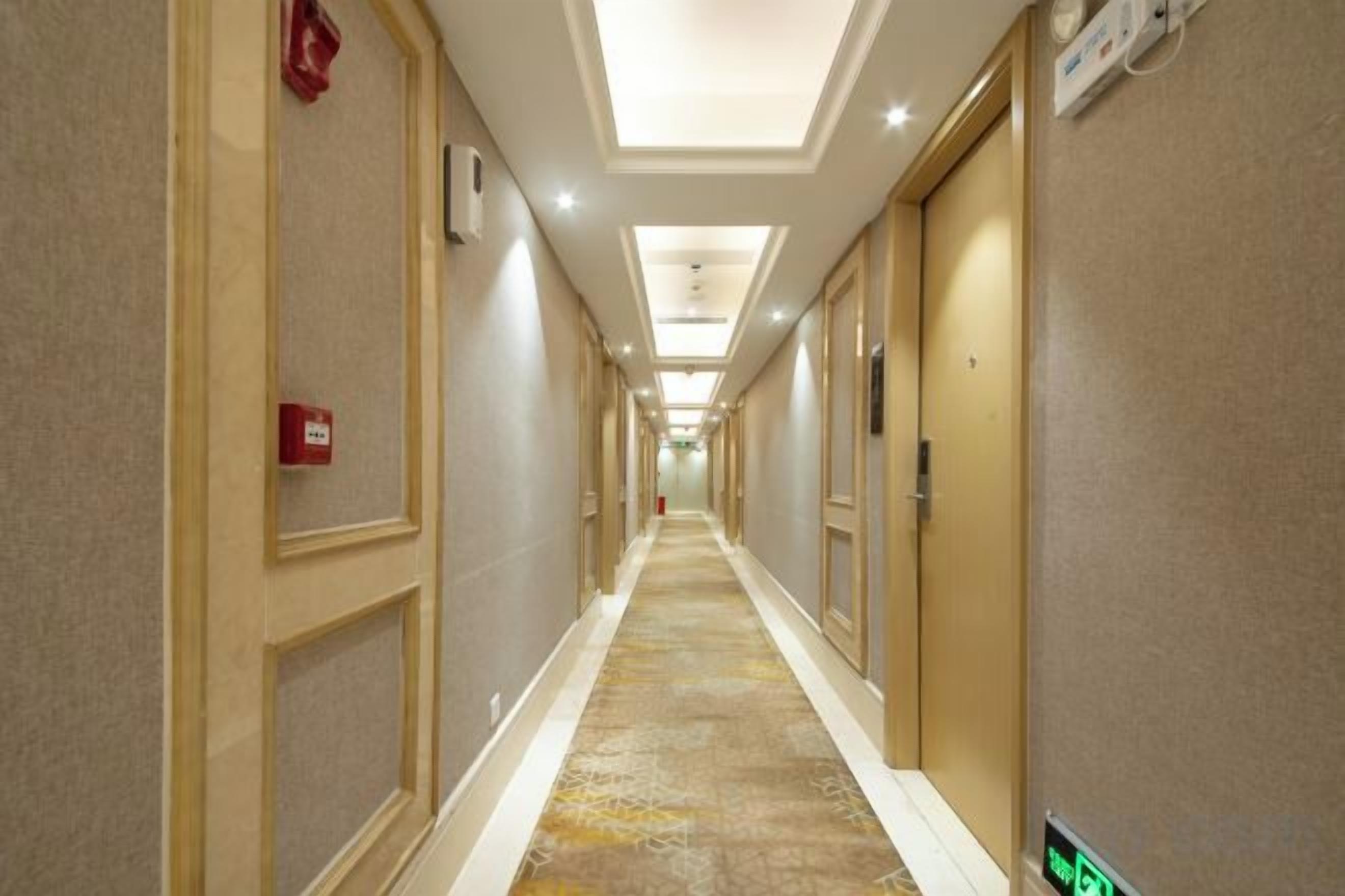 A hotel corridor with doors on both sides, a carpeted floor, and a fire alarm on the left wall.