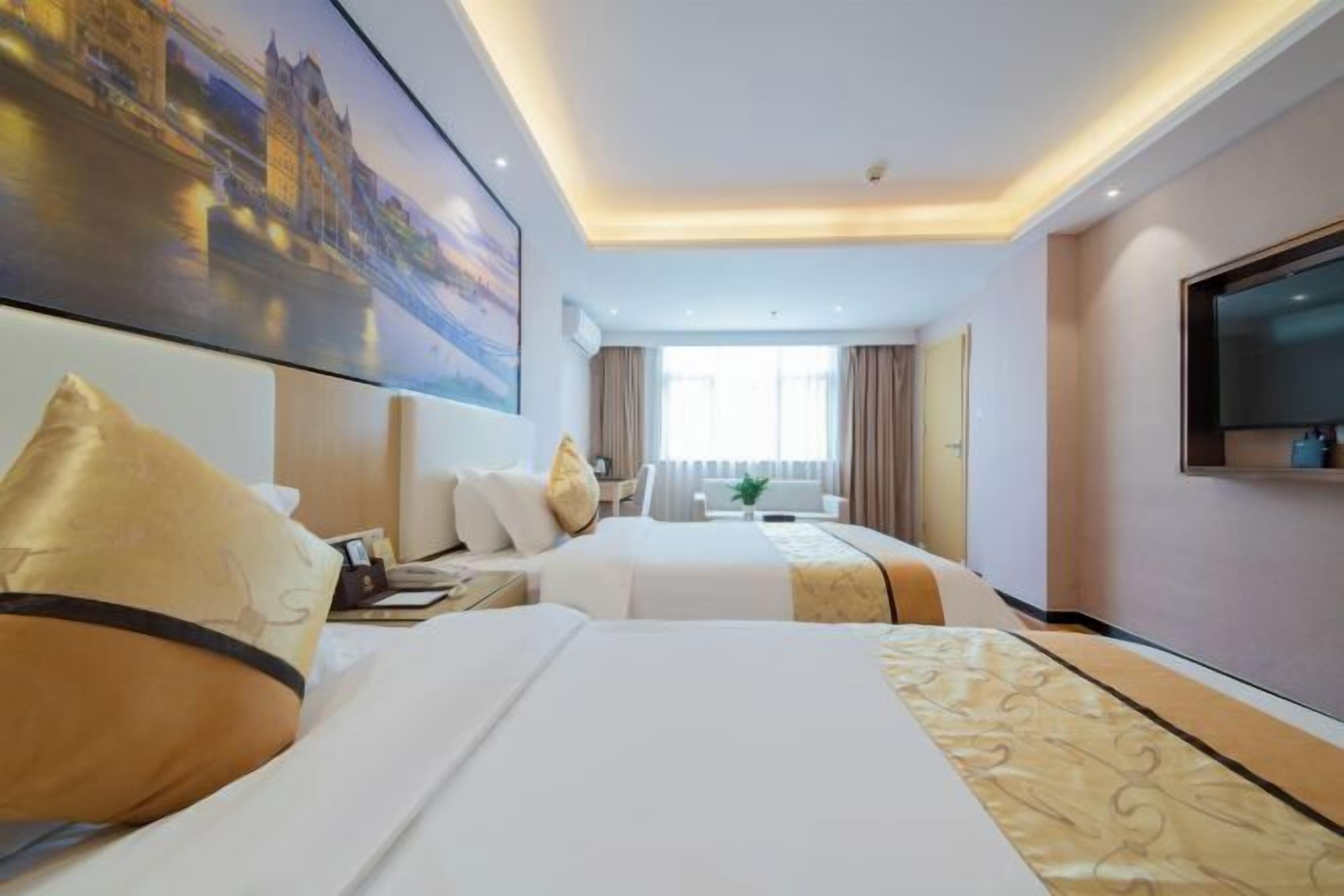 Une chambre d’hôtel avec deux lits, un grand téléviseur à écran plat, un tableau représentant un paysage urbain et une fenêtre avec des rideaux.