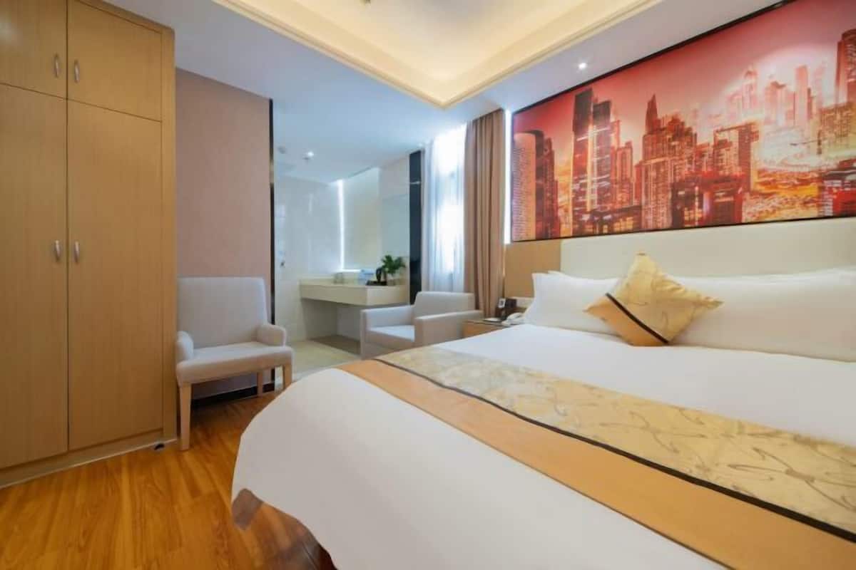 A hotel room with a large bed, a seating area, a wardrobe, and a cityscape painting on the wall.