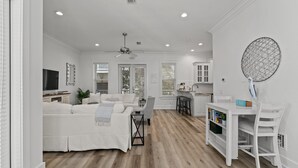A spacious living area with a white sofa, a dining table, and a kitchen area with a white cabinet and a round decorative item on the wall. - Gorgeous, spacious home, steps to the 12,000sqft pool, recently renovated. Perfect for the whole family! (Panama City Beach)