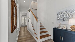 A hallway with a wooden staircase, a dark blue cabinet with a decorative item, and a wall with a metallic art piece. - Gorgeous, spacious home, steps to the 12,000sqft pool, recently renovated. Perfect for the whole family! (Panama City Beach)