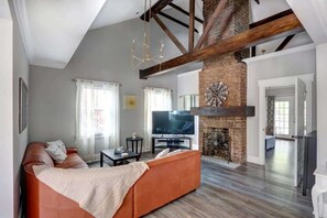 Amplia sala de estar con techo alto, vigas de madera a la vista, chimenea de ladrillo, televisor plano grande y sofá naranja confortable.