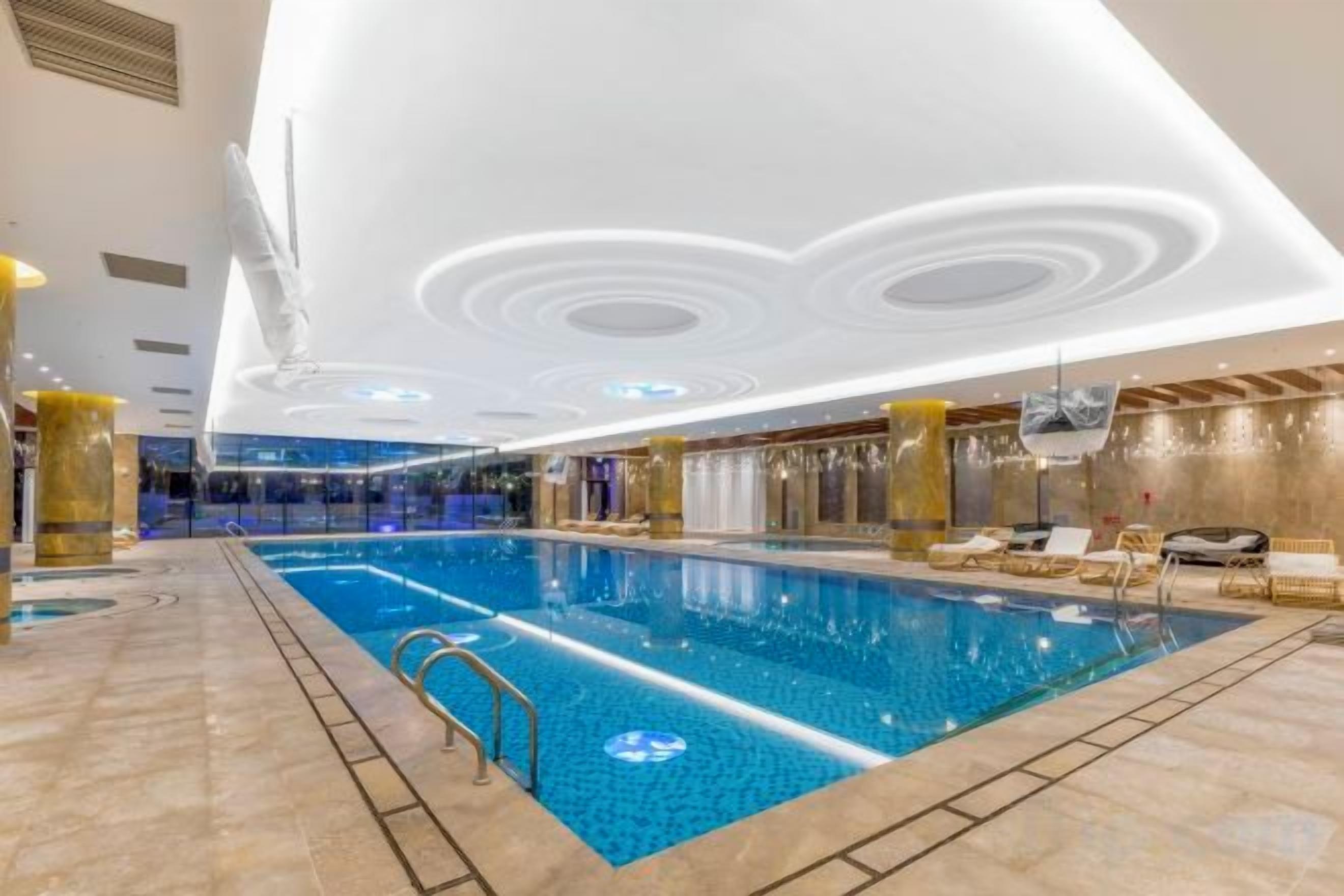 An indoor swimming pool with a tiled floor, a pool ladder, and a ceiling with circular light fixtures.
