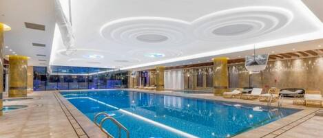 An indoor swimming pool with a tiled floor, a pool ladder, and a ceiling with circular light fixtures.