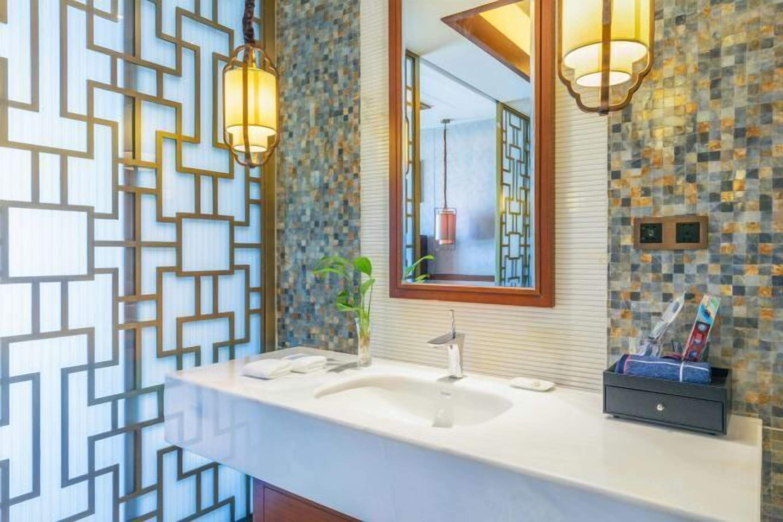 Une salle de bain avec un lavabo blanc, deux appliques murales, un miroir avec un cadre en bois et un mur décoratif orné d’un motif géométrique.