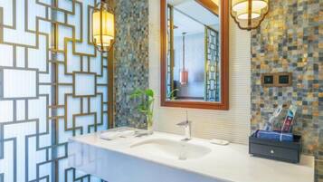 Une salle de bain avec un lavabo blanc, deux appliques murales, un miroir avec un cadre en bois et un mur décoratif orné d’un motif géométrique.