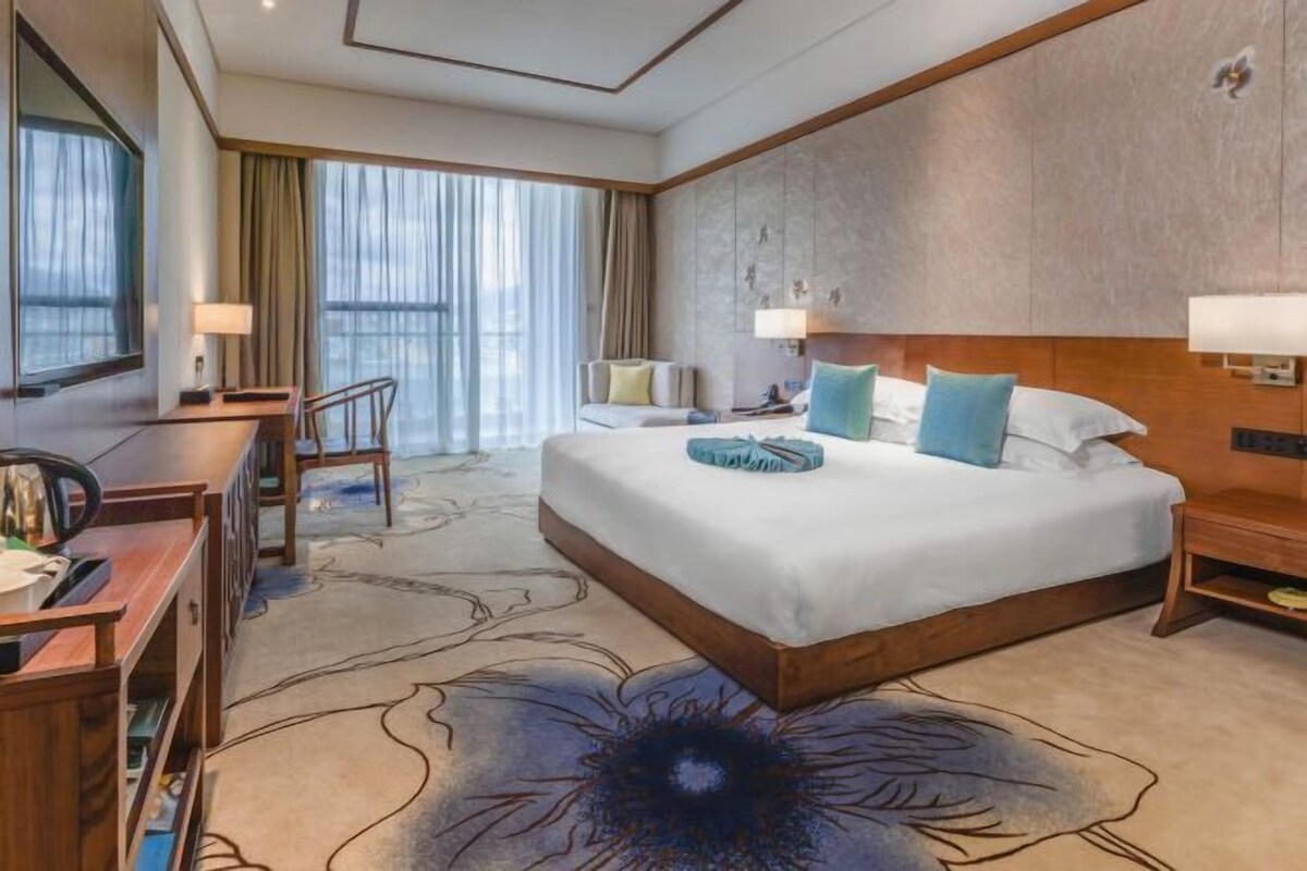 Habitación de hotel con una cama grande, un escritorio, una silla y vistas a la ciudad a través del ventanal.