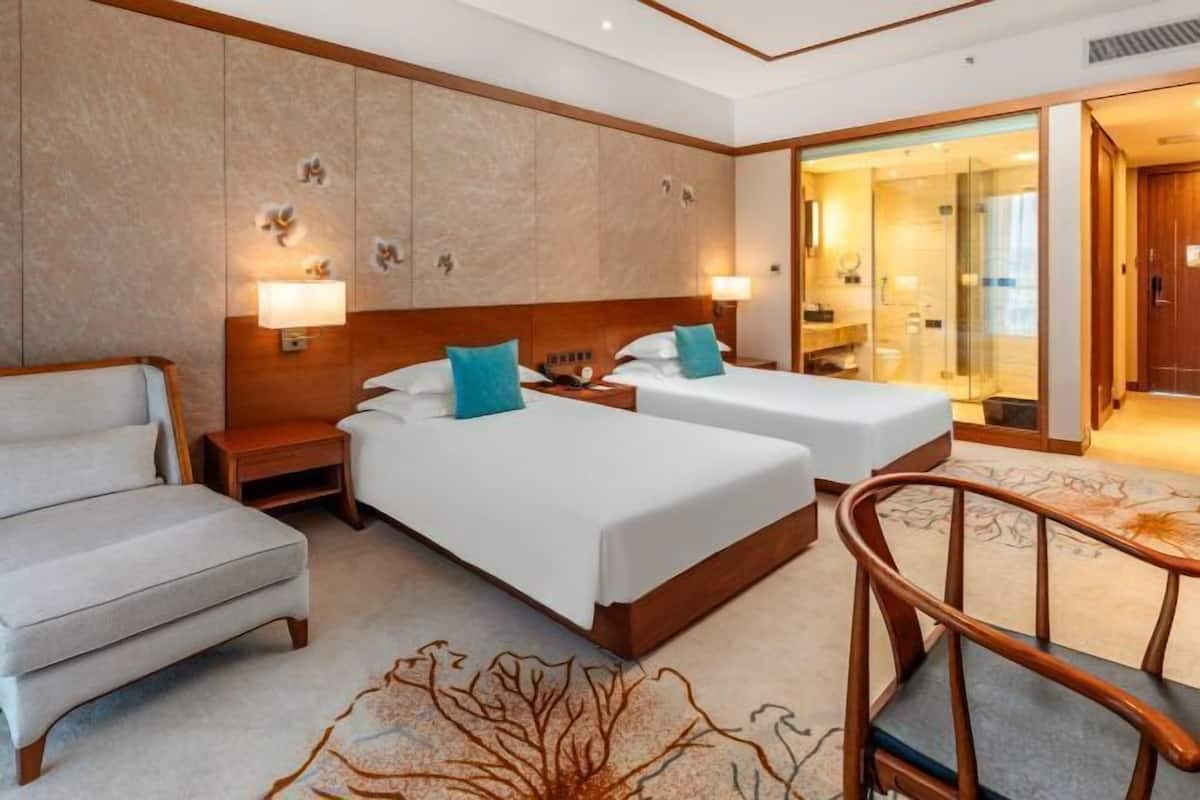 Une chambre d’hôtel avec deux lits, un canapé, une chaise en bois et une salle de bain visible à travers une porte en verre.