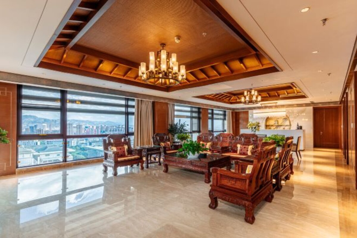 A spacious lobby with a high ceiling, large windows offering a city view, and a central seating area with wooden furniture.