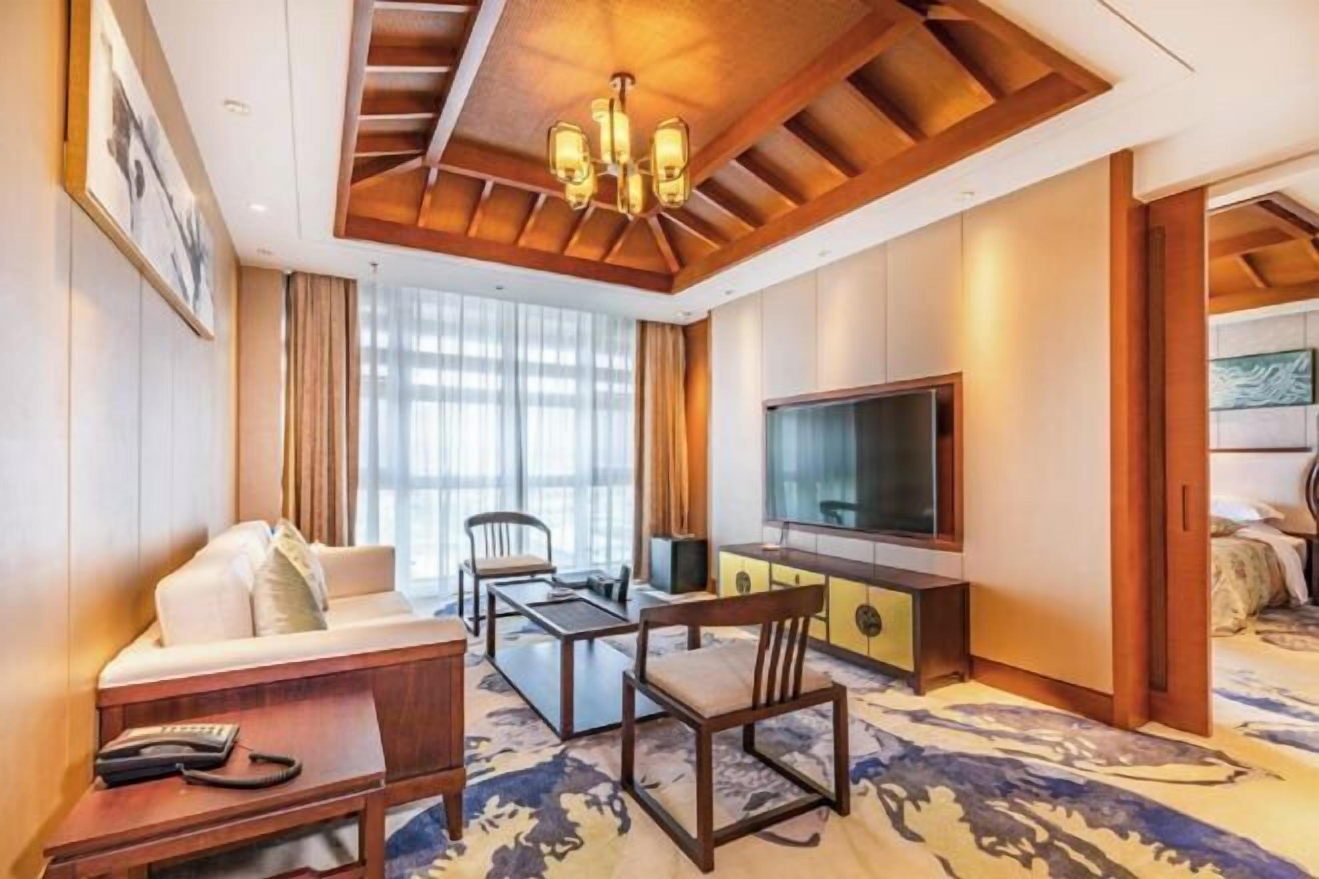 A hotel room with a sofa, a TV, a desk, and a chair. There is a window with curtains and a chandelier on the ceiling.
