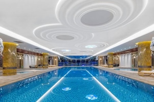 A modern indoor swimming pool with a central lane, surrounded by seating areas and a decorative ceiling.