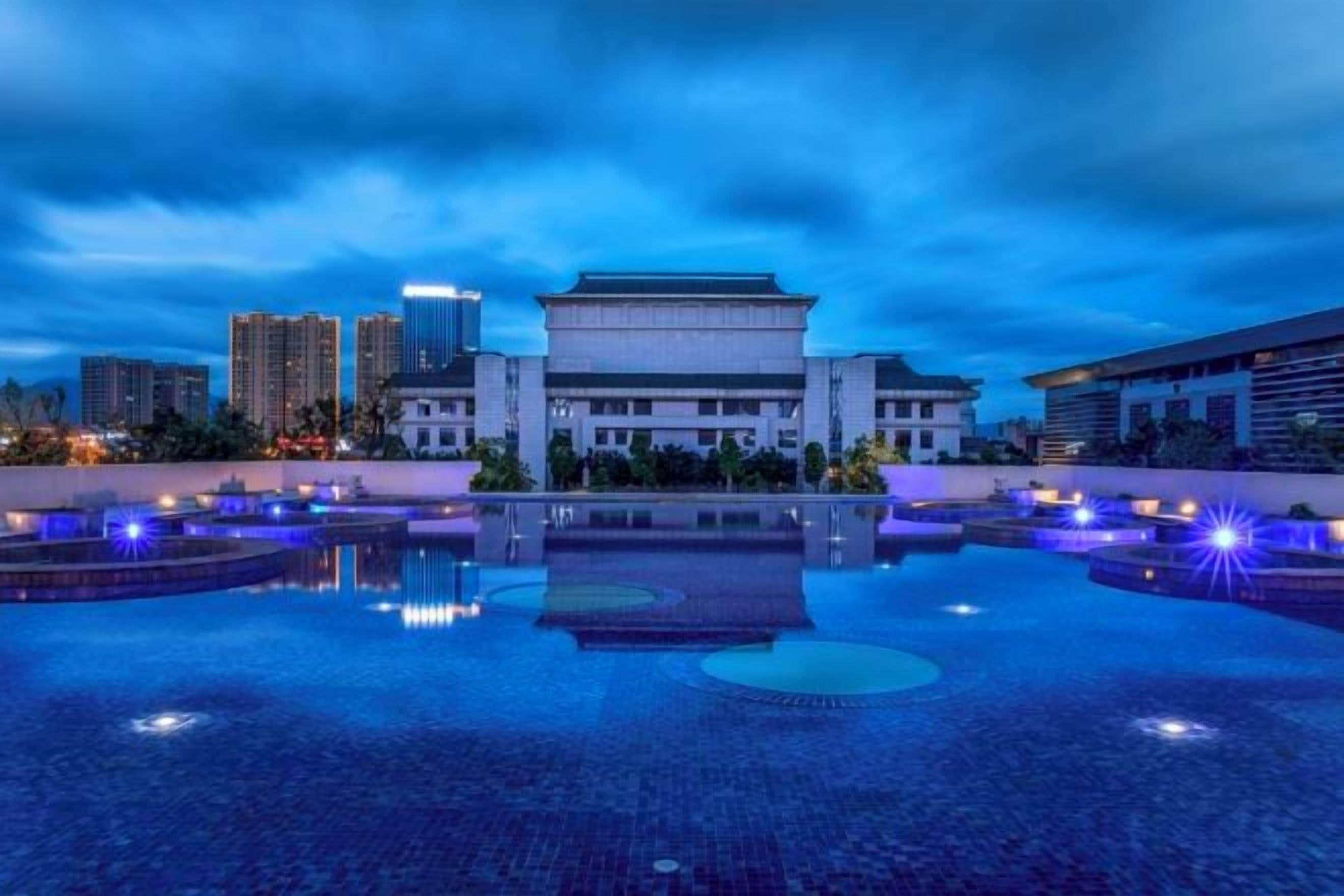 A building with a traditional architectural style, a reflective water feature in front, and modern high-rise buildings in the background.