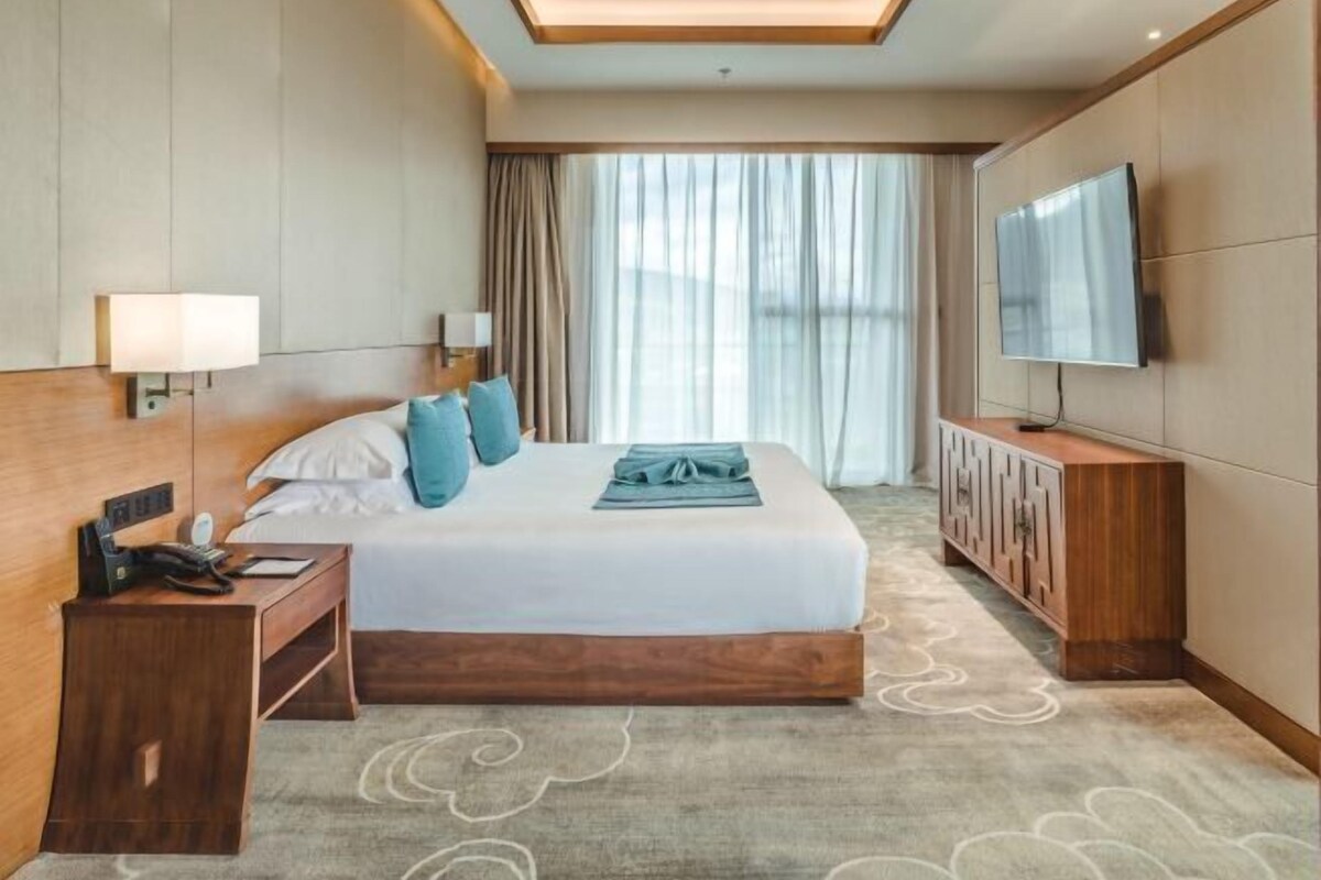 Habitación de hotel con cama, mesita de noche, lámpara, televisor y vistas al exterior.