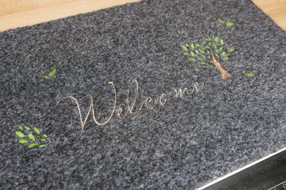 A dark textured mat with 'Welcome' embroidered in gold and small green leaf designs.
