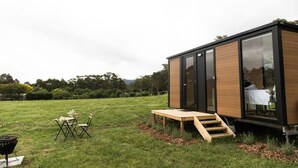 A modern, compact structure with large windows, a wooden deck, and a small outdoor seating area.