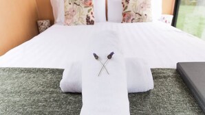 A neatly made bed with floral pillows, a white towel, and two lavender flowers.