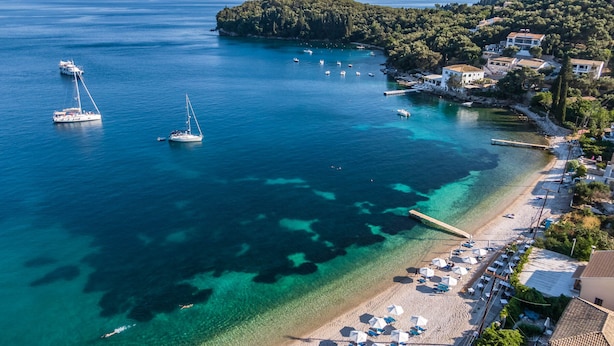 An aerial view of a coastal area with clear blue waters, boats, and a beach with umbrellas and sun loungers. - Kalami Beach - Villa Mara (Corfu)