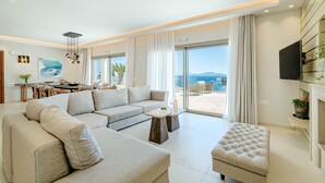 A modern living room with a large sectional sofa, a small ottoman, and a dining area with a table and chairs. - Kalami Beach - Villa Mara (Corfu)