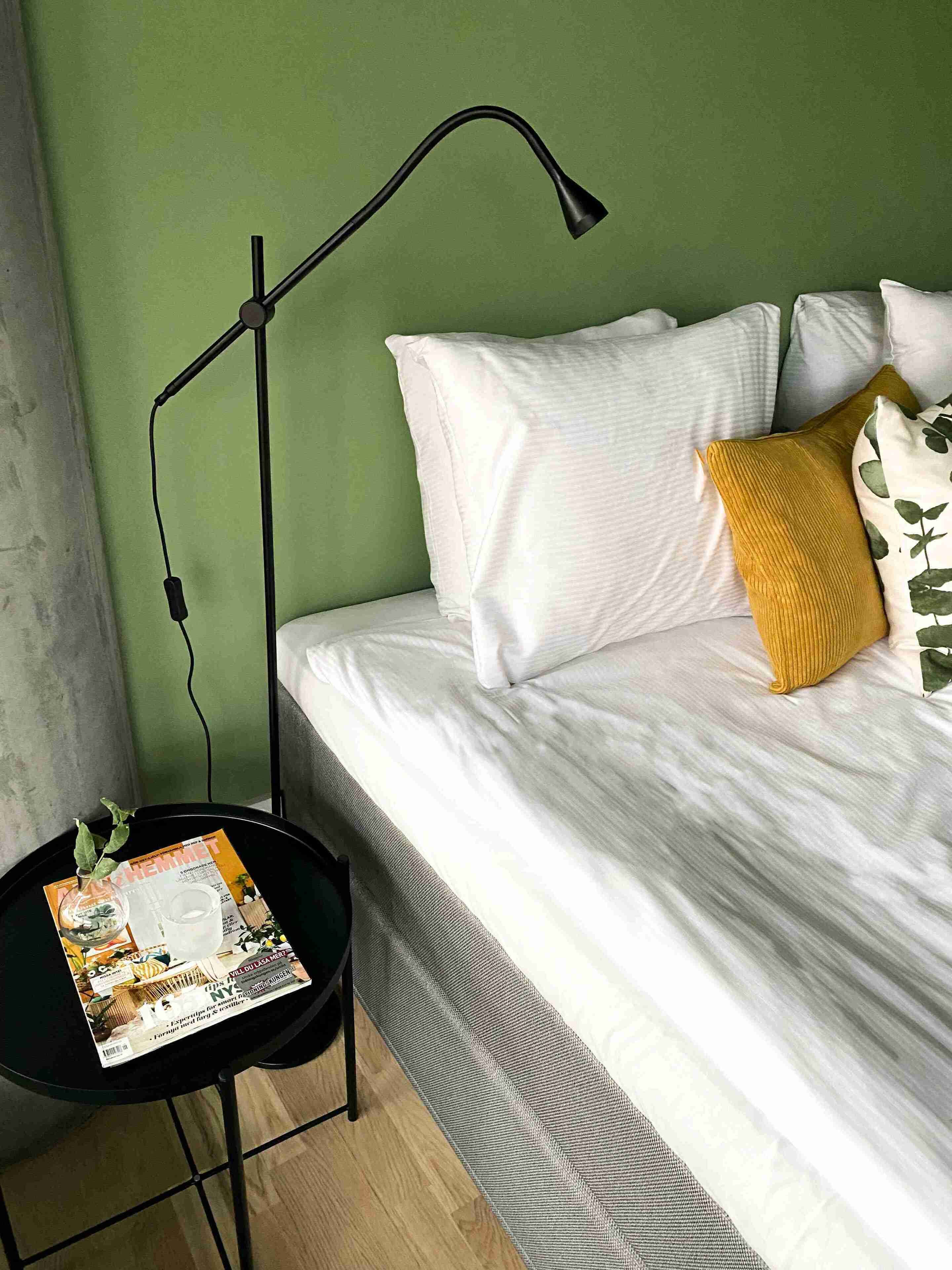 Una cama bien hecha con almohadas blancas y amarillas, una lámpara de pie negra y una revista sobre una mesita redonda al lado de la cama.
