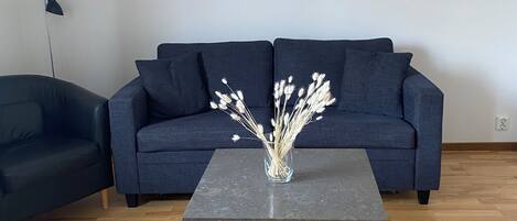 Una sala de estar moderna con un sofá azul oscuro, una mesa de centro de vidrio con estructura metálica y un florero con flores blancas.