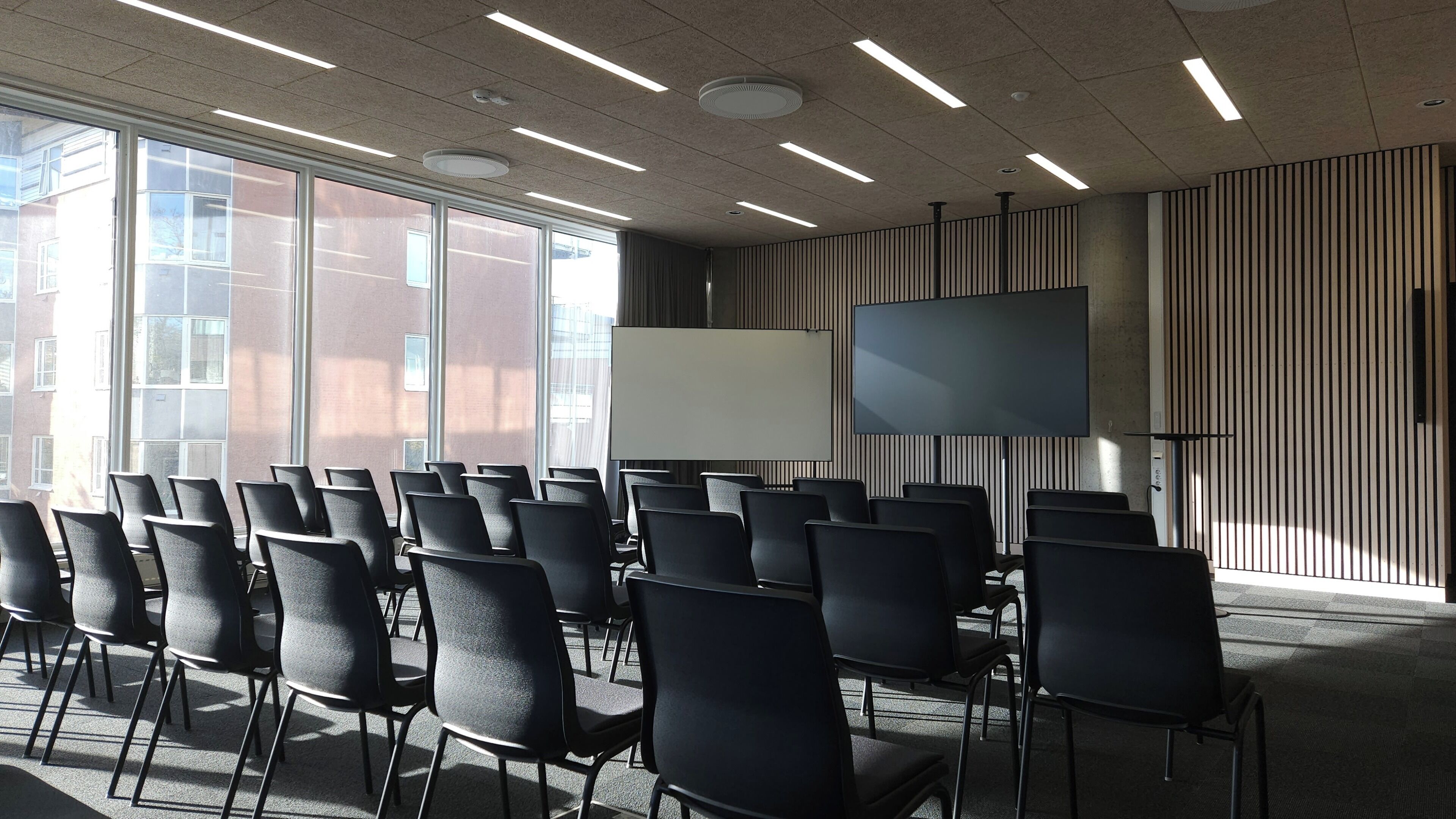 A conference room with rows of chairs, a large screen, and a view of buildings through large windows.