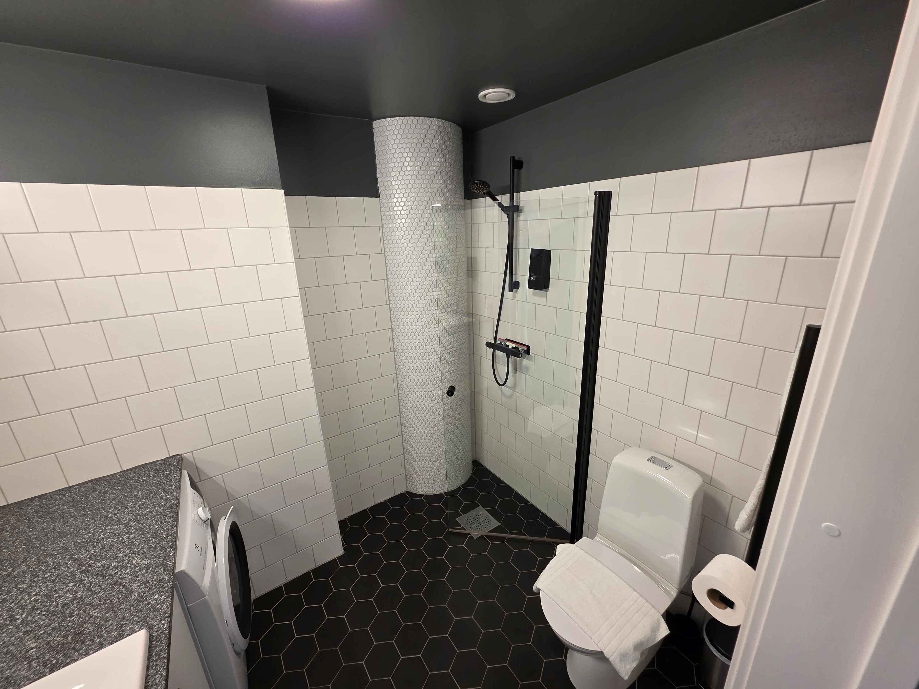 Un baño con baldosas hexagonales blanco y negro, una ducha con puerta de cristal y un inodoro con una toalla.