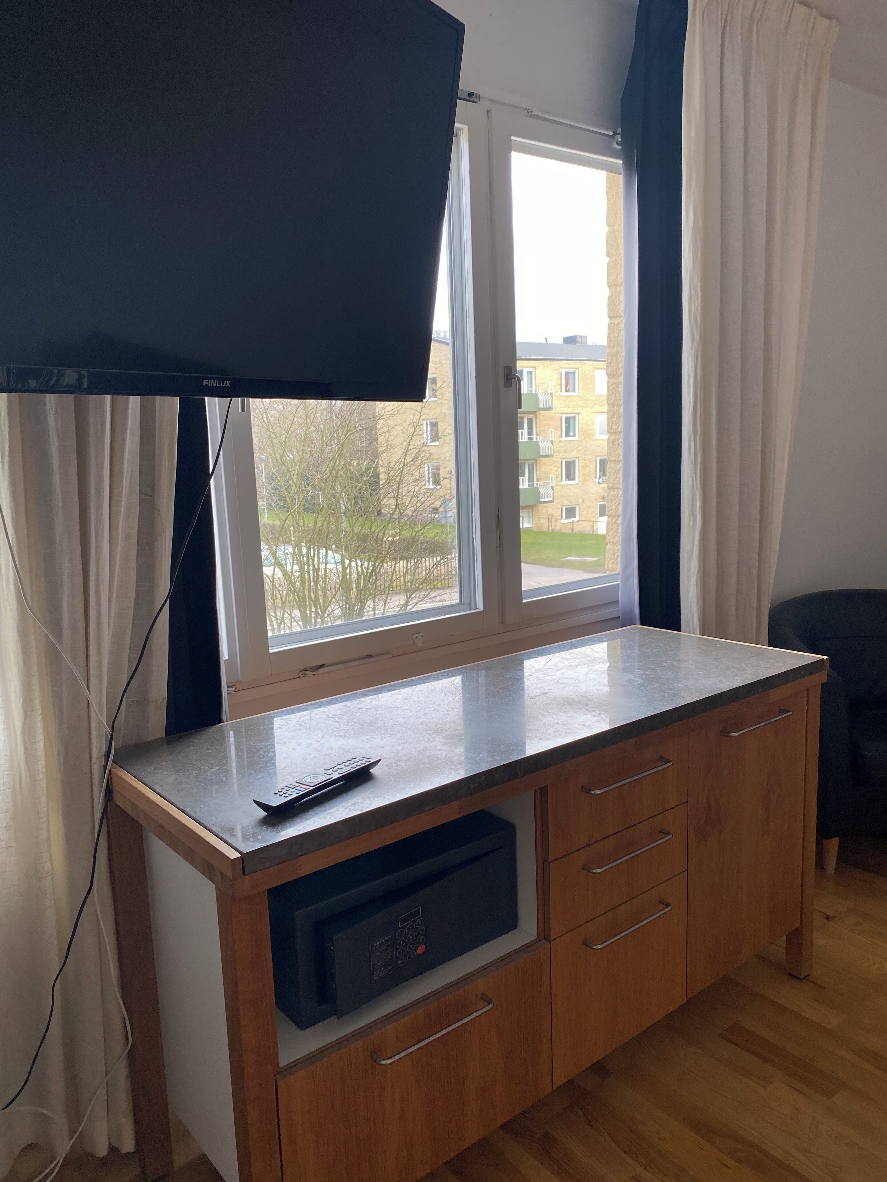 A room with a flat-screen TV mounted on the wall, a wooden cabinet with a built-in microwave, and a window with curtains.