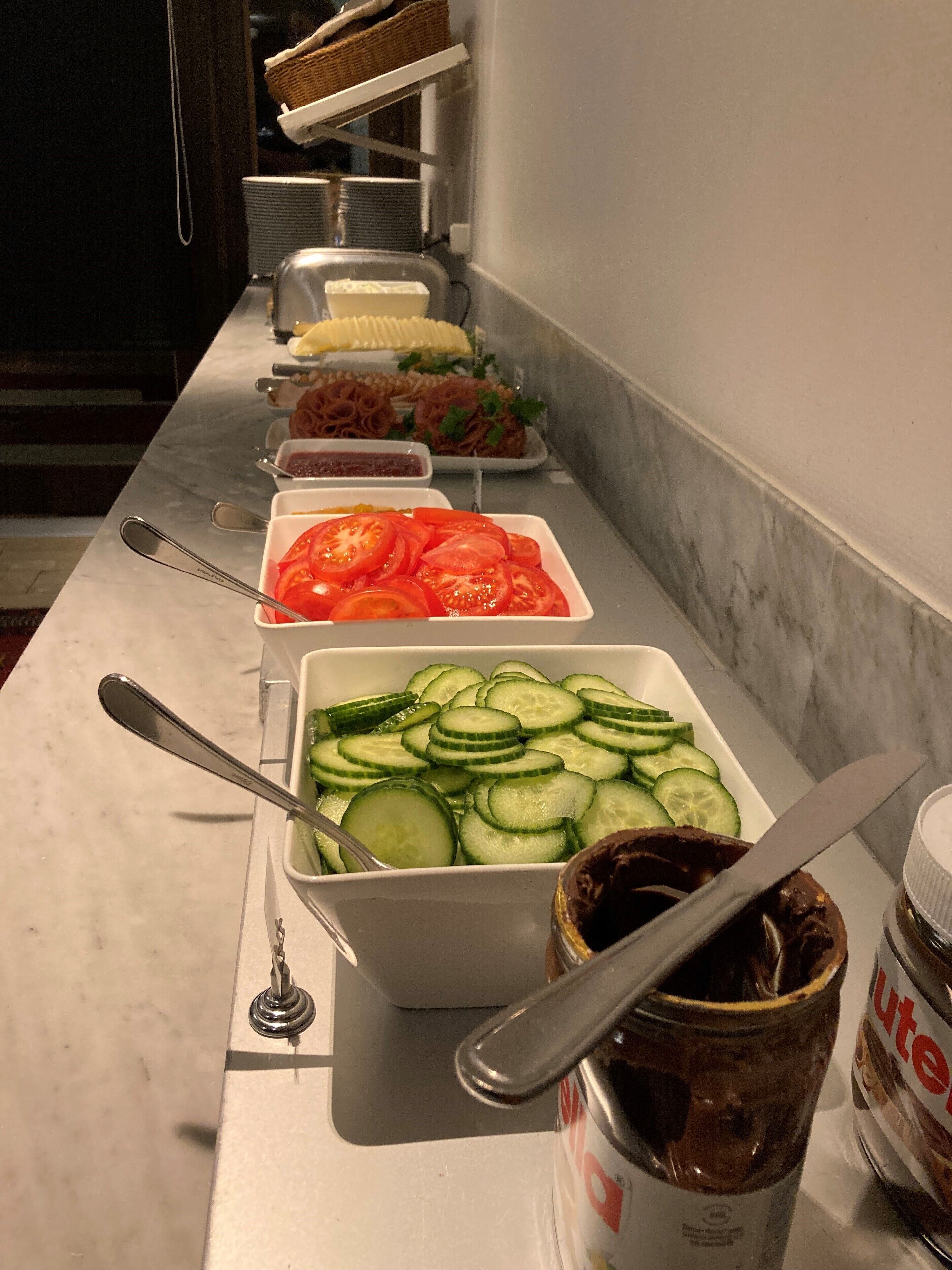Una mesa de buffet con diferentes alimentos, incluyendo tomates en rodajas, pepinos y un tarro de Nutella.