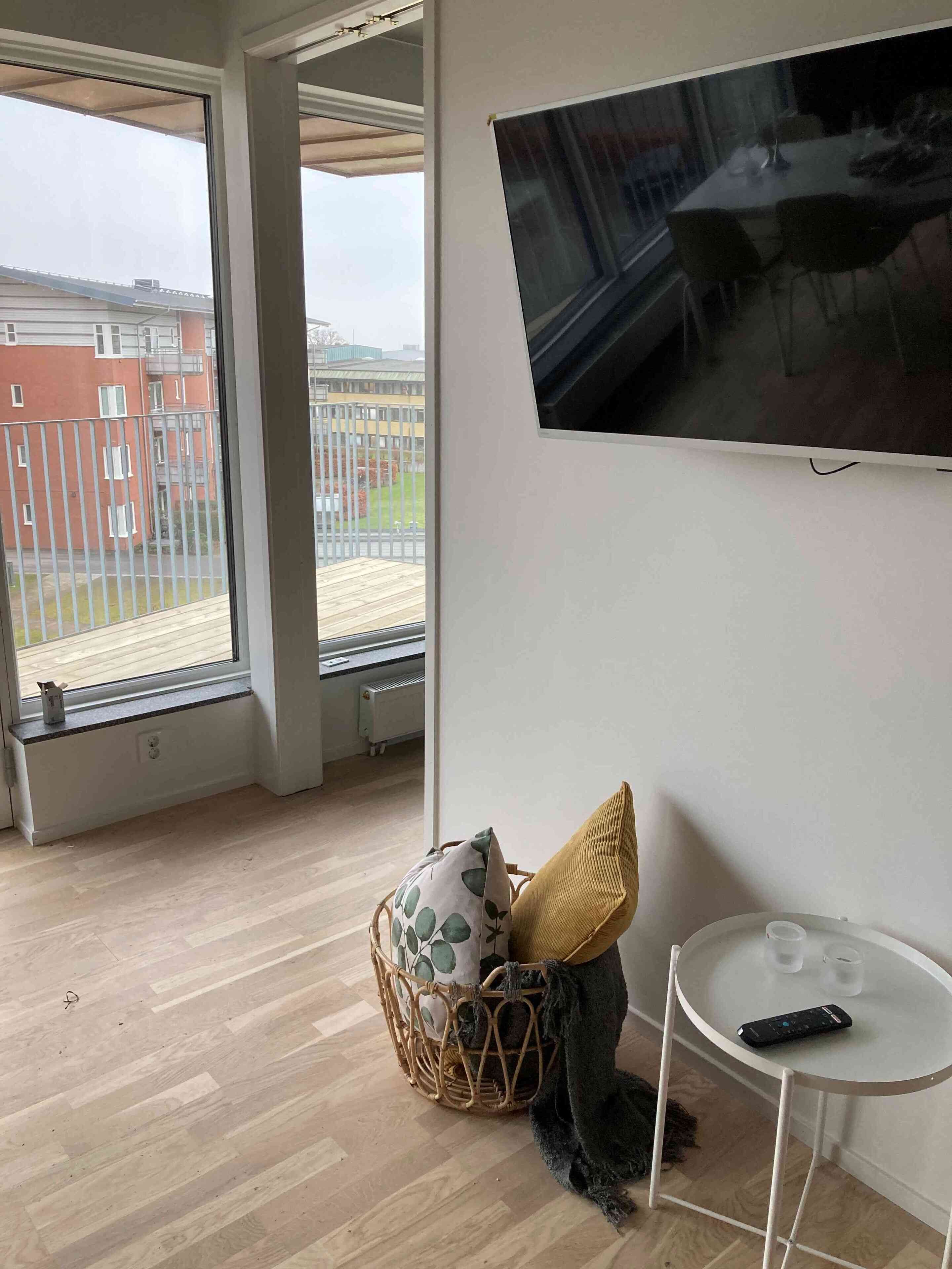 A modern room with a large window, a flat-screen TV mounted on the wall, a small round table, and a wicker chair with cushions.